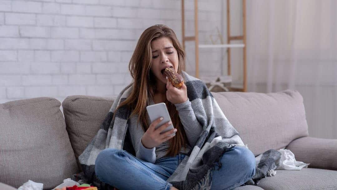 Woman sitting on a sofa eating comfort food during an emotional eating moment