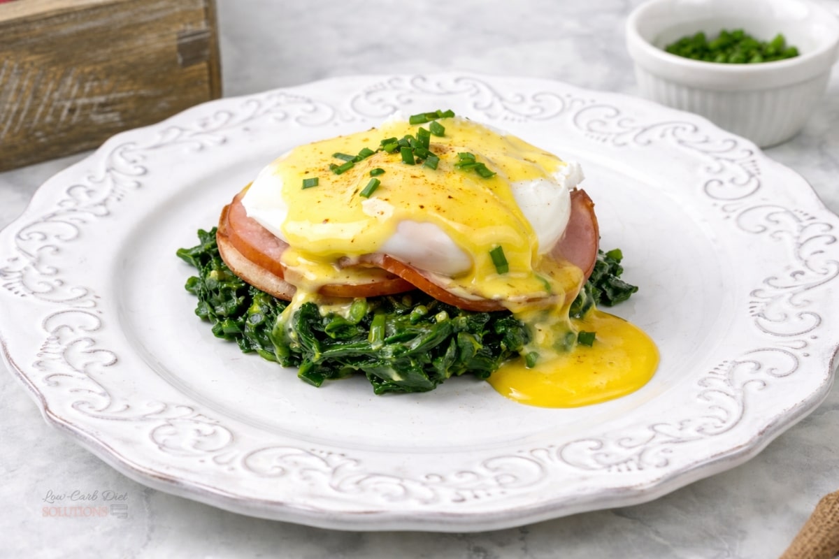 Keto Eggs Benedict with sautéed spinach, Canadian bacon, and a poached egg topped with hollandaise, garnished with chopped chives and a pinch of cayenne pepper