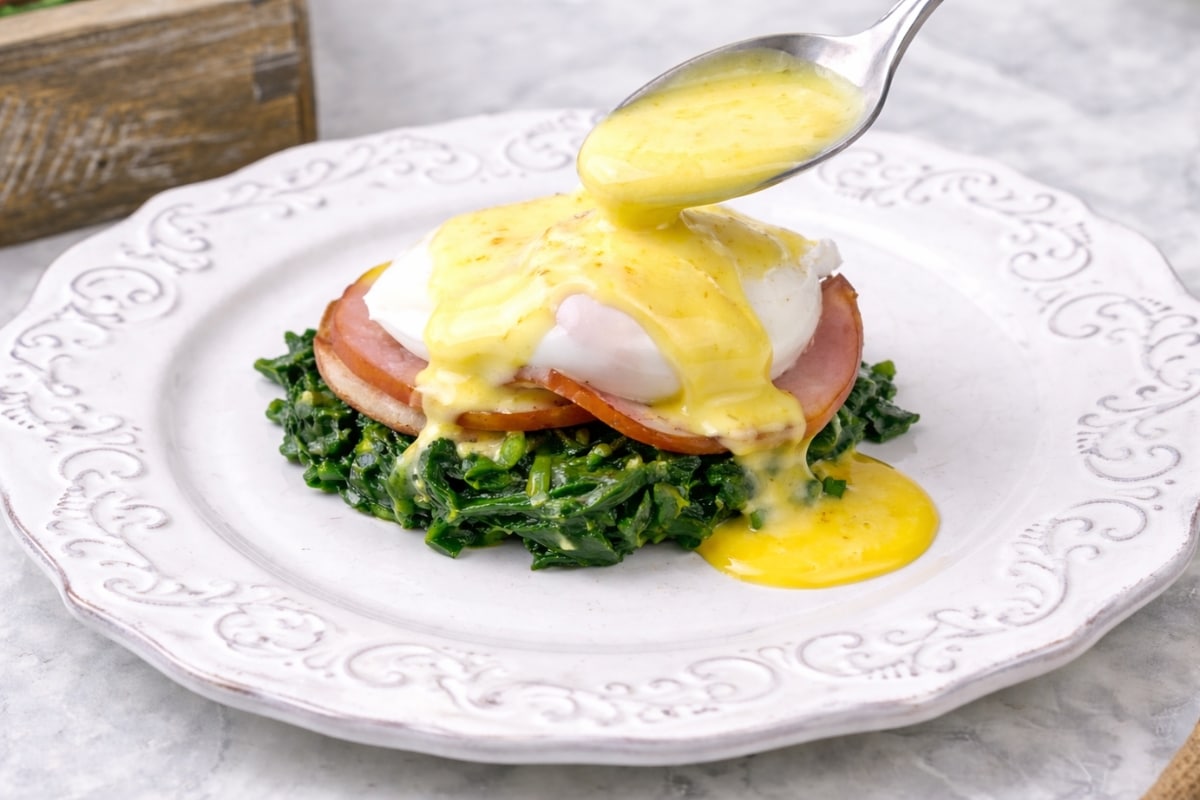 Spooning warm hollandaise sauce over a poached egg with Canadian bacon and sautéed spinach