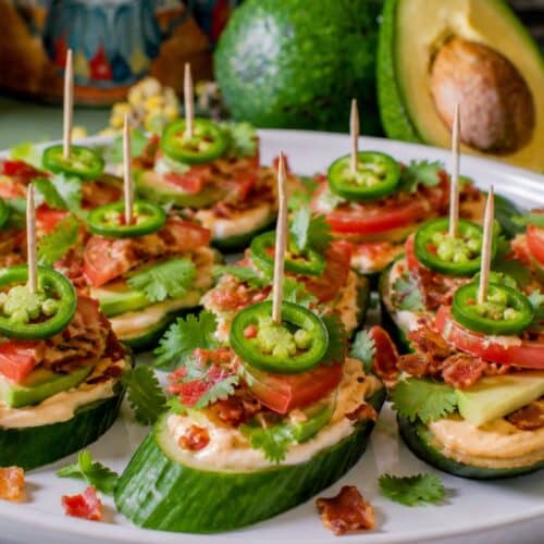 keto cucumber bites topped with avocado tomato cilantro and visible jalapeno slices on a white appetizer platter