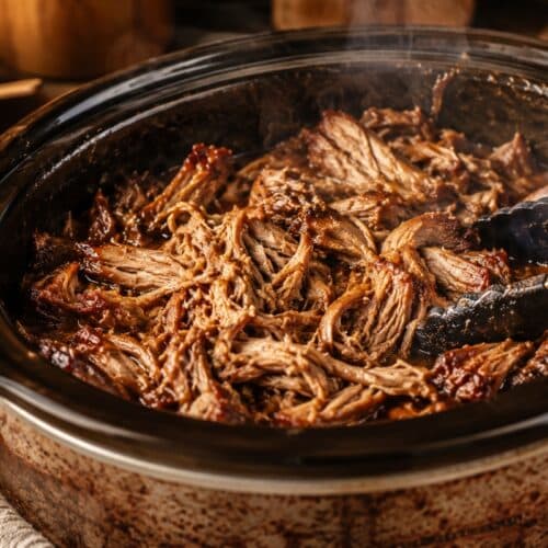 Slow-cooker pulled pork inside a rustic crockpot with tender shredded pork, rich cooking juices, caramelised edges, steam rising, and serving tongs in a warm cozy kitchen setting